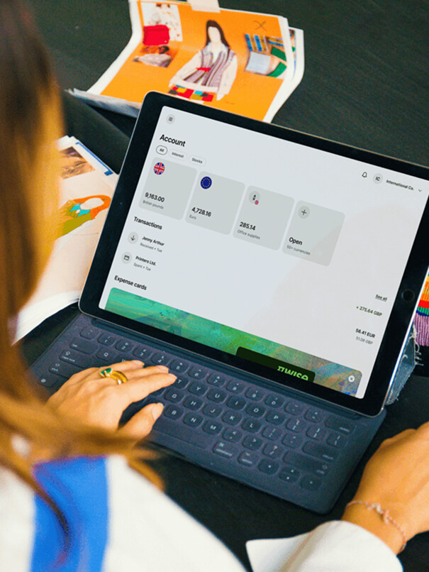 Une personne, vue de dos par-dessus son épaule, est en train de taper sur une tablette. L'écran de la tablette affiche un tableau de bord intitulé « Compte » avec les balances, les transactions et les « Cartes de dépenses ».