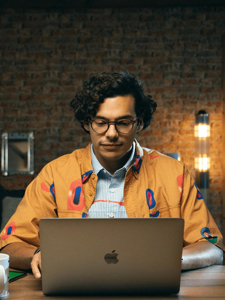Un homme portant des lunettes et une veste orange distinctive est assis à un bureau, penché sur un ordinateur portable. En arrière-plan se trouve un mur en briques. Les mots « POUR LES PETITES ENTREPRISES » apparaissent en blanc en haut de l'image, tandis qu'un cercle vert avec un signe « + » blanc est visible en bas à droite.
